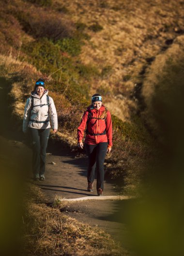 Zwei Wanderinnen begleiten sich auf einem Pfad in einer hügeligen, natürlichen Landschaft bei sonnigem Wetter. | © Kleinwalsertal Tourismus eGen • Fotograf: Hannah Bichay