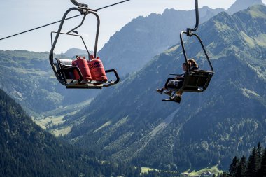 Two people sitting in a summer ski lift above a green alpine village surrounded by mountains. | © Kleinwalsertal Tourismus eGen • Photographer: Oliver Farys