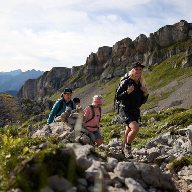Four hikers climbing a rocky mountain trail in Gottesacker in Kleinwalsertal with mountain peaks in the background on a sunny day. | © Kleinwalsertal Tourismus eGen • Photographer: Oliver Farys