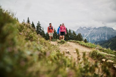 Three hikers on a mountain trail in Kleinwalsertal, surrounded by green meadows and mountains under a cloudy sky. | © Kleinwalsertal Tourismus eGen • Photographer: Oliver Farys