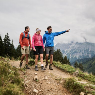 Four hikers, two women and two men, walking on a mountain trail in the mountains, with one man pointing forward. | © Kleinwalsertal Tourismus eGen • Photographer: Oliver Farys