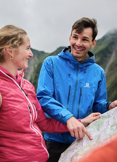 Zwei Wanderer mit Rucksack, lachend und sich unterhaltend während einer Herbstwanderung im Kleinwalsertal, mit Bergpanorama im Hintergrund. | © Kleinwalsertal Tourismus eGen • Fotograf: Oliver Farys