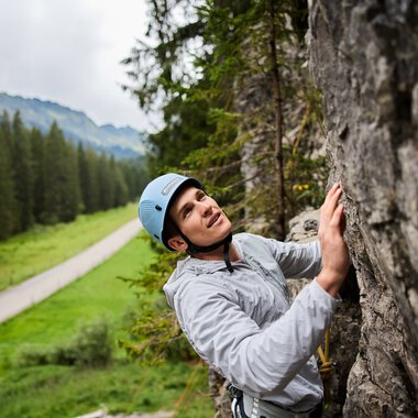 Woman climbing at a rock climbing area in the mountains, surrounded by green forests and a path in the background. | © Kleinwalsertal Tourismus eGen • Photographer: Oliver Farys