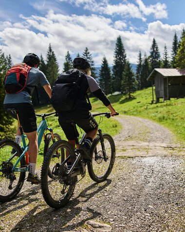 Two mountain bikers on a forest trail in Kleinwalsertal, surrounded by trees and a small hut in the background, sunny sky. | © Kleinwalsertal Tourismus eGen • Photographer: Oliver Farys