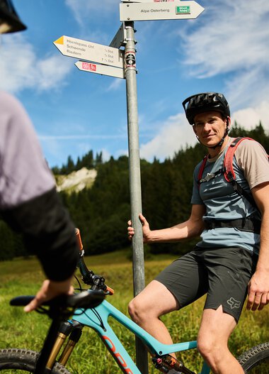 Ein Mann beim Mountainbiken hält an einem Wegweiser in einer grünen, bergigen Landschaft bei sommerlichem Himmel. | © Kleinwalsertal Tourismus eGen • Fotograf: Oliver Farys