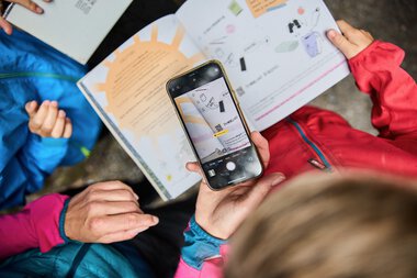 Kinder beim Naturforscherabenteuer am Schwarzwasserbach im Kleinwalsertal, eines fotografiert mit einem Smartphone, während das andere ein Informationsblatt hält. | © Kleinwalsertal Tourismus eGen • Fotograf: Oliver Farys