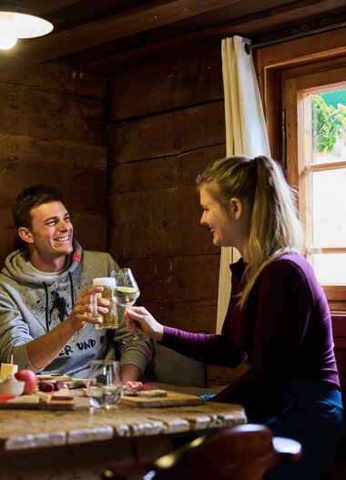 Zwei Personen sitzen in einer Holzunterkunft, lachen und stoßen sich mit Weingläsern an, während sie drinnen essen. | © Kleinwalsertal Tourismus eGen • Fotograf: Oliver Farys
