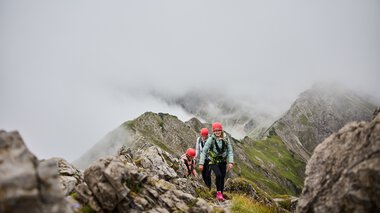 Menschen beim Klettersteig in den Bergen, umgeben von Wolken und Felsen, mit Bergschule Walter Walser-Logo im Bild. | © Kleinwalsertal Tourismus eGen • Fotograf: Oliver Farys