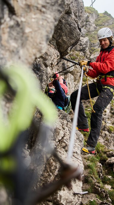 Women rock climbing on a via ferrata in the mountains, wearing harnesses, natural scenery in the background.
