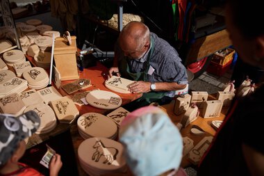 An elderly man signing or painting wooden items at a market stall in Kleinwalsertal. | © Kleinwalsertal Tourismus eGen • Photographer: Oliver Farys