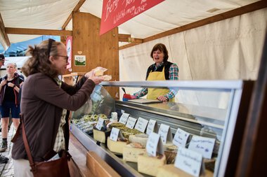 Photo of a cheese stand at a weekly market in Kleinwalsertal with a vendor selling cheese to a customer. | © Kleinwalsertal Tourismus eGen • Photographer: Oliver Farys