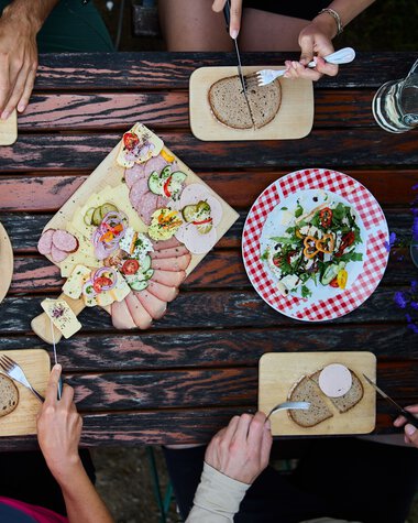 Blick von oben auf einen Tisch mit verschiedenen Käse- und Wurstplatten, Brot, und Salat, umgeben von vier Personen beim Essen. | © Kleinwalsertal Tourismus eGen • Fotograf: Oliver Farys