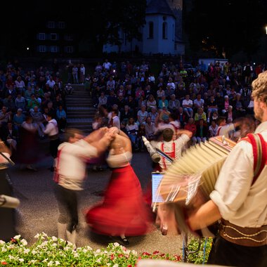 Musicians in traditional attire perform at a summer festival in Kleinwalsertal as a large crowd dances and watches in the background. | © Kleinwalsertal Tourismus eGen • Photographer: Oliver Farys