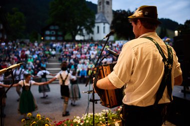 Musician on stage at summer festival in Kleinwalsertal, front view of a man with hat playing an accordion, with a dance group and crowd near a church in the background. | © Kleinwalsertal Tourismus eGen • Photographer: Oliver Farys