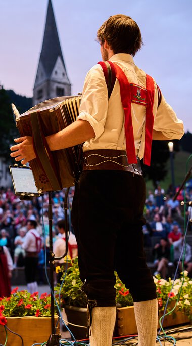 Musiker auf einer Bühne beim Alpenzauber-Festival im Kleinwalsertal, spielt Gitarre während die Menge im Hintergrund zuschaut, mit Bergkulisse und Kirche im Hintergrund. | © Kleinwalsertal Tourismus eGen • Fotograf: Oliver Farys