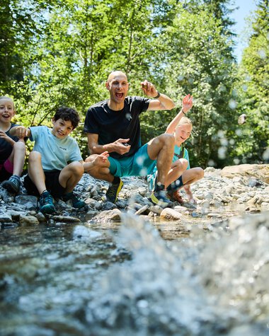A lively group of four people, including children and an adult, sitting by a creek in a green forest, enjoying playing in the water. | © Kleinwalsertal Tourismus eGen • Photographer: Oliver Farys