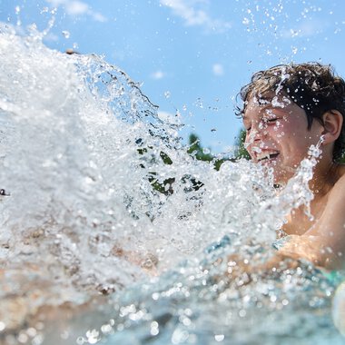 A happy woman splashes in an outdoor pool on a sunny day, water splashing around her. | © Kleinwalsertal Tourismus eGen • Photographer: Oliver Farys