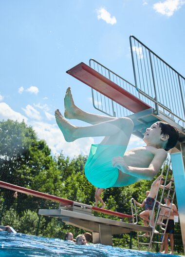 Ein Junge springt vom Sprungbrett in das Freibad im Sommer, mit anderen Badegästen und grünen Bergen im Hintergrund. | © Kleinwalsertal Tourismus eGen • Fotograf: Oliver Farys