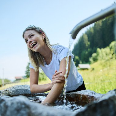 Frau sitzt am Kneippbecken, lacht und nutzt die kalte Wassertherapie im Grünen bei schönem Wetter. | © Kleinwalsertal Tourismus eGen • Fotograf: Oliver Farys