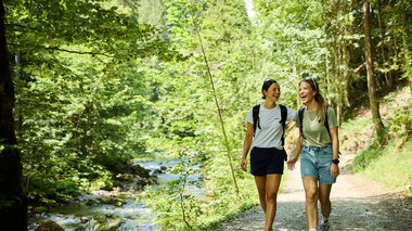 Two young women walking and laughing along a lush forest trail by a river during summer 2024, surrounded by nature and sunshine. | © Kleinwalsertal Tourismus eGen • Photographer: Oliver Farys
