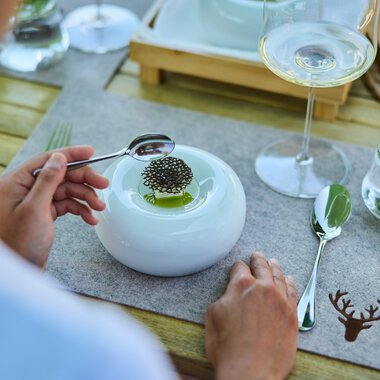 A table setting featuring a modern white plate with a small dish and chopsticks, a glass of white wine, a water glass with mint leaves, and utensils on a wooden table with a nature-themed tablecloth. | © Kleinwalsertal Tourismus eGen • Photographer: Oliver Farys