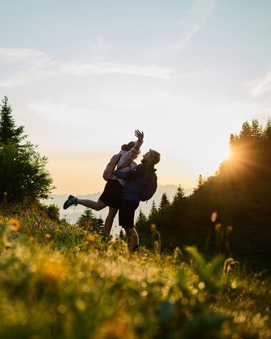 Ein Paar hebt sich bei Sonnenaufgang in einer grünen Bergwiese im Kleinwalsertal hoch, umgeben von Bäumen, mit Sonnenlicht im Hintergrund. | © Kleinwalsertal Tourismus eGen • Fotograf: Oliver Farys