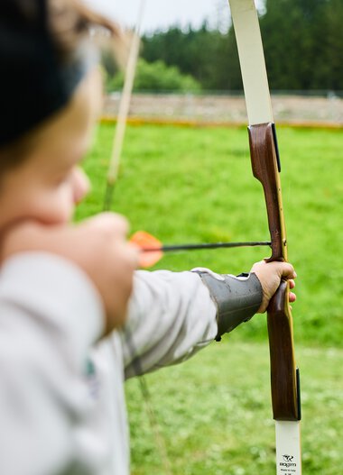 A child practicing archery at a wilderness school in Kleinwalsertal, aiming at a target in the background, surrounded by green nature. | © Kleinwalsertal Tourismus eGen • Photographer: Oliver Farys