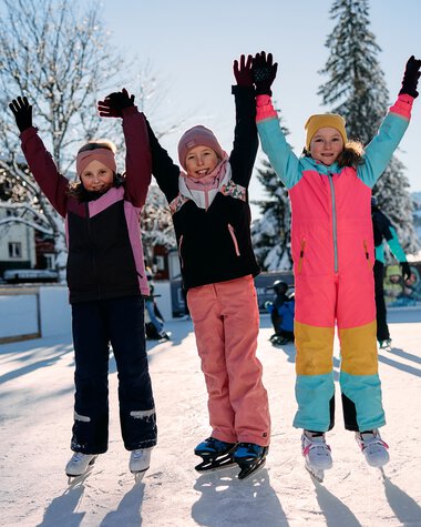Children ice skating on a frozen lake in winter, joyfully raising their arms, with snow-covered trees and other skaters around. | © Kleinwalsertal Tourismus eGen • Photographer: Oliver Farys
