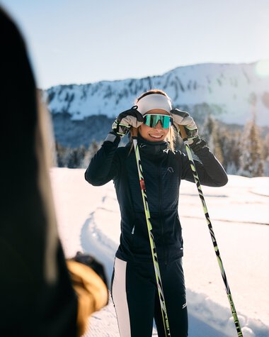Woman cross-country skiing in the snow, wearing a ski helmet and holding ski poles, with snow-covered mountains in the background during sunrise or sunset. | © Kleinwalsertal Tourismus eGen • Photographer: Oliver Farys