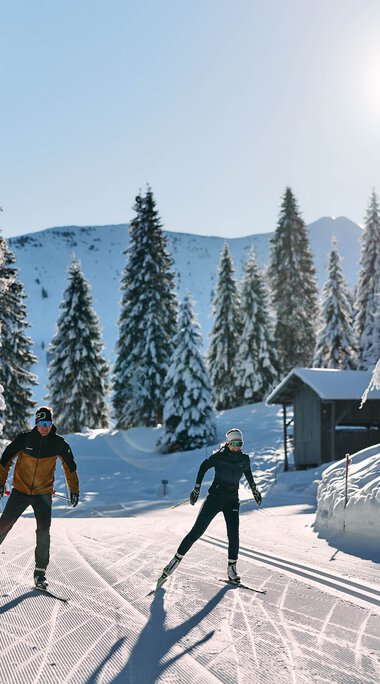 Two cross-country skiers enjoying skiing in a snowy winter forest in Kleinwalsertal under sunny skies. | © Kleinwalsertal Tourismus eGen • Photographer: Oliver Farys