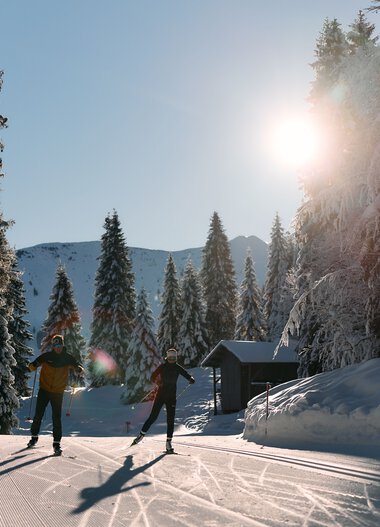 Two cross-country skiers in a snowy winter landscape under bright sunshine in Kleinwalsertal. | © Kleinwalsertal Tourismus eGen • Photographer: Oliver Farys