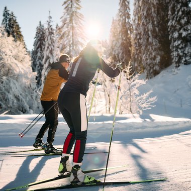 Two cross-country skiers in a snowy winter landscape, sunlight filtering through trees, alpine scenery in the background. | © Kleinwalsertal Tourismus eGen • Photographer: Oliver Farys