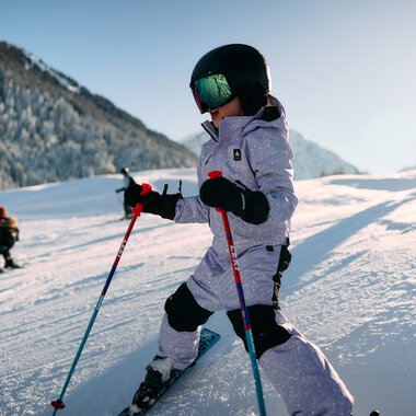 Child skiing in the snowy landscape, sunlight, mountains in the background, wearing helmet and ski gear. | © Kleinwalsertal Tourismus eGen • Photographer: Oliver Farys