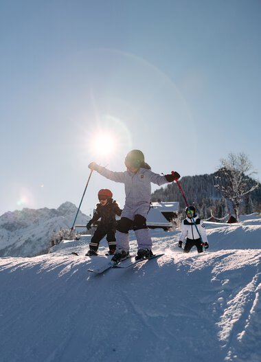 Kinder beim Skifahren auf einer schneebedeckten Piste in den Bergen bei Sonnenlicht, begleitet von einem Erwachsenen. | © Kleinwalsertal Tourismus eGen • Fotograf: Oliver Farys