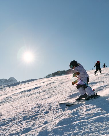 Kleines Kind beim Skifahren auf einer schneebedeckten Piste, sonniger Tag, andere Kinder im Hintergrund, Berg im Hintergrund. | © Kleinwalsertal Tourismus eGen • Fotograf: Oliver Farys