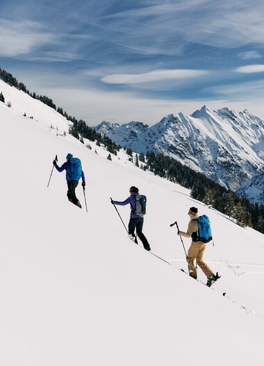 Drei Skitourengeher klettern an einer verschneiten Berghänge im Kleinwalsertal mit Bergen im Hintergrund. | © Kleinwalsertal Tourismus eGen • Fotograf: Oliver Farys