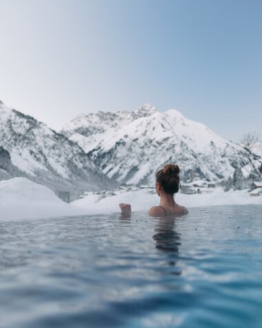 Person entspannt im warmen Pool inmitten verschneiter Berge und Winterlandschaft im Kleinwalsertal. | © Kleinwalsertal Tourismus eGen • Fotograf: Oliver Farys
