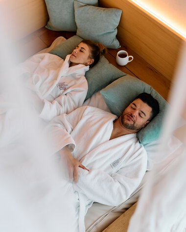A man and a woman are lying on lounge beds in a wellness spa area, wrapped in bathrobes, with pillows and a cup. They look relaxed and calm. | © Kleinwalsertal Tourismus eGen • Photographer: Oliver Farys