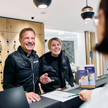 Two smiling people at the reception during the Winter 2025 event in Kleinwalsertal, behind the counter at the tourism office. | © Kleinwalsertal Tourismus eGen • Photographer: Oliver Farys