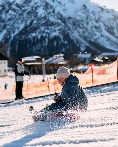 A child sleds in the snow at Kleinwalsertal, surrounded by mountains and other winter sports enthusiasts on a sunny day. | © Kleinwalsertal Tourismus eGen • Photographer: Oliver Farys