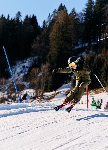 Children skiing on a snow-covered slope in Kleinwalsertal, surrounded by trees and winter scenery, in sunny weather. | © Kleinwalsertal Tourismus eGen • Photographer: Oliver Farys
