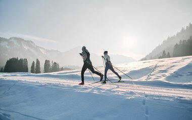 Two cross-country skiers skiing on the snowy Schwarzwasserloipe trail in Kleinwalsertal during sunrise, surrounded by mountains and pine trees. | © Kleinwalsertal Tourismus eGen • Photographer: Oliver Farys