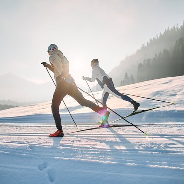 Zwei Menschen beim Langlaufen auf der Schwarzwasserloipe im Kleinwalsertal, umgeben von schneebedeckter Landschaft und Bergen im Hintergrund, bei Sonnenaufgang. | ©  Kleinwalsertal Tourismus eGen • Fotograf: Oliver Farys