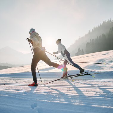 Zwei Skifahrer beim Langlaufen auf der Schwarzwasserloipe im Kleinwalsertal, umgeben von Bergen und Wald, bei Sonnenaufgang. | ©  Kleinwalsertal Tourismus eGen • Fotograf: Oliver Farys