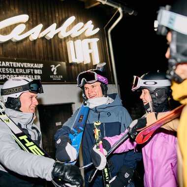 Four skiers in winter gear and helmets, laughing and chatting at night in a ski resort, with illuminated sign 'Kaiser Lift' and a signboard in the background. | © Kleinwalsertal Tourismus eGen • Photographer: Oliver Farys