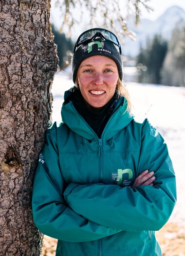 Portrait of a smiling woman in winter clothing, with arms crossed, standing next to a tree with snow and mountains in the background. | © Kleinwalsertal Tourismus eGen • Photographer: Oliver Farys
