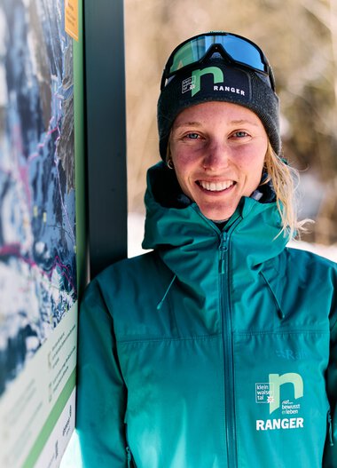 A smiling woman in winter gear with ski goggles stands next to an information sign in the snow, with trees and a snowy landscape in the background. | © Kleinwalsertal Tourismus eGen • Photographer: Oliver Farys