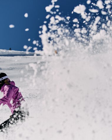Skifahrerin in pink beim Abfahrtsrennen im Schnee, mit Sprühspur und weiter Berg im Hintergrund. | © Kleinwalsertal Tourismus eGen • Fotograf: Oliver Farys