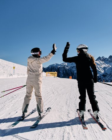 Zweimal Skifahrer geben sich auf einer schneebedeckten Piste beim Skifahren in den Bergen ein High-Five, im Hintergrund die Alpen im klaren blauen Himmel. | © Kleinwalsertal Tourismus eGen • Fotograf: Oliver Farys