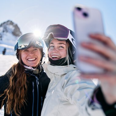 Zwei lachende Wintersportlerinnen beim Skifahren auf der Kanzelwand im Kleinwalsertal, die ein Selfie mit dem Handy machen. | ©  Kleinwalsertal Tourismus eGen • Fotograf: Oliver Farys